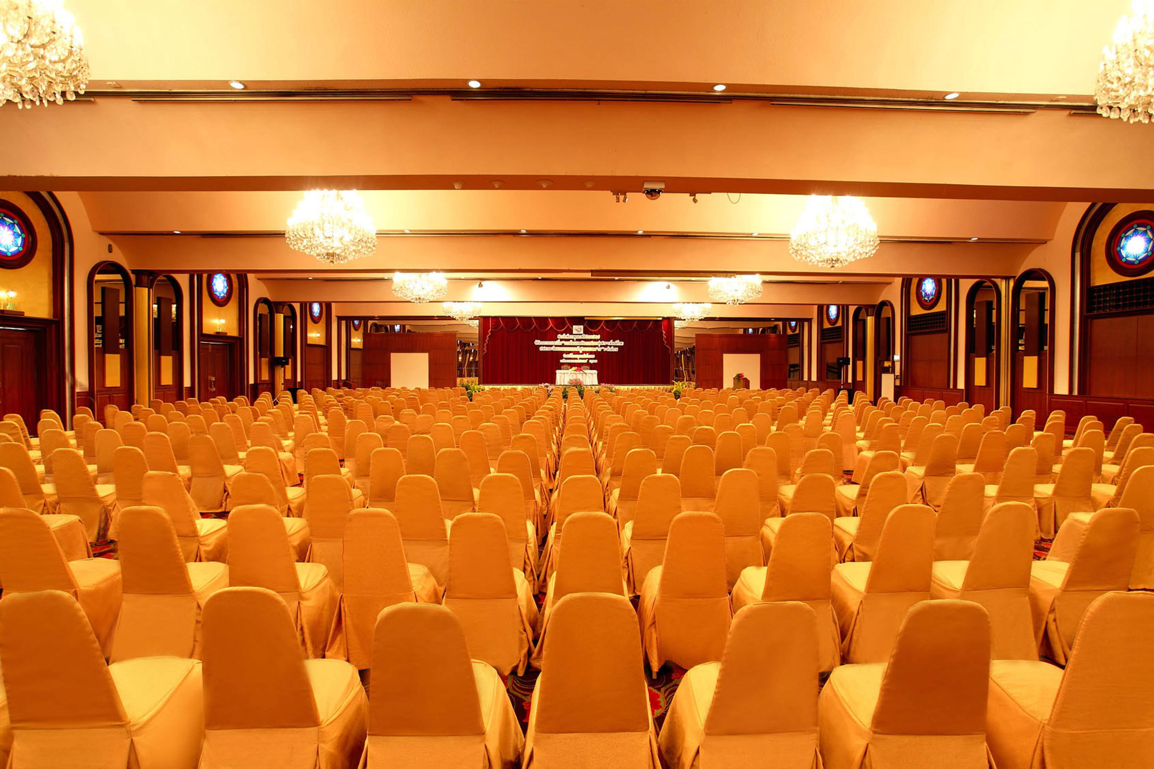 Een ruime congreszaal met rijen stoelen, een podium met een rode achtergrond en kroonluchters.