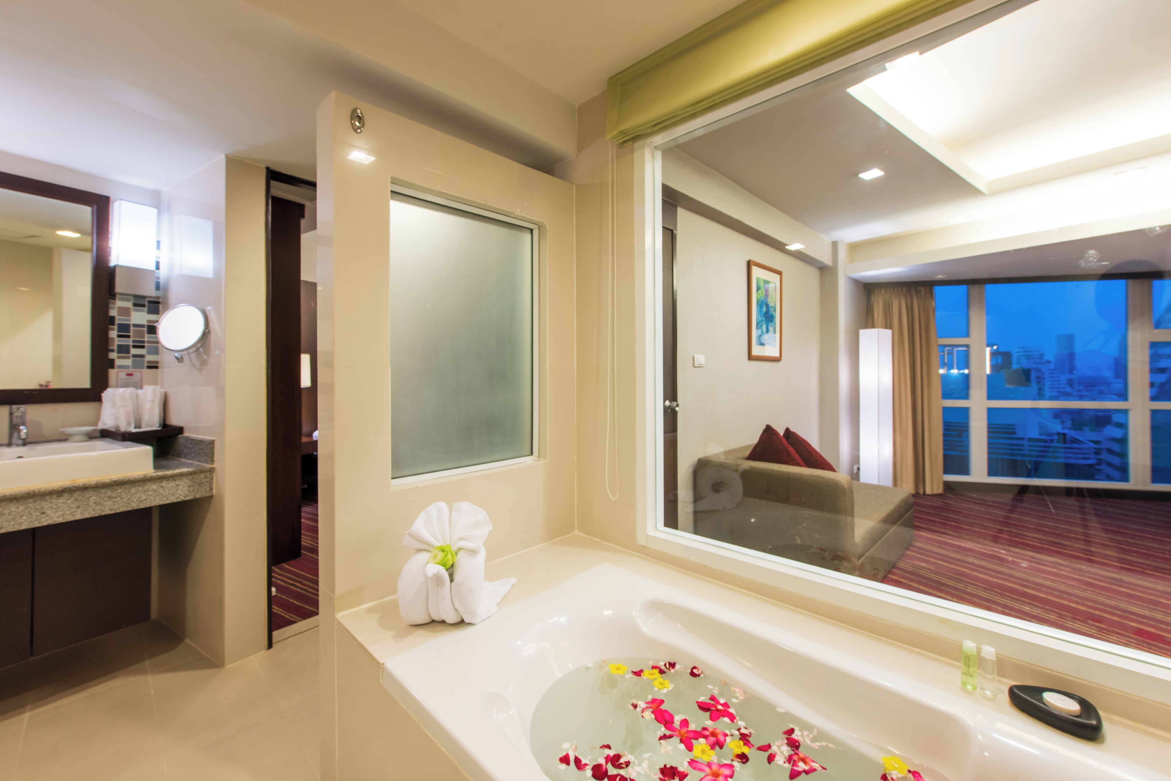 A hotel bathroom with a large mirror, a sink, a bathtub with floral arrangements, and a view of a cityscape through a large window.