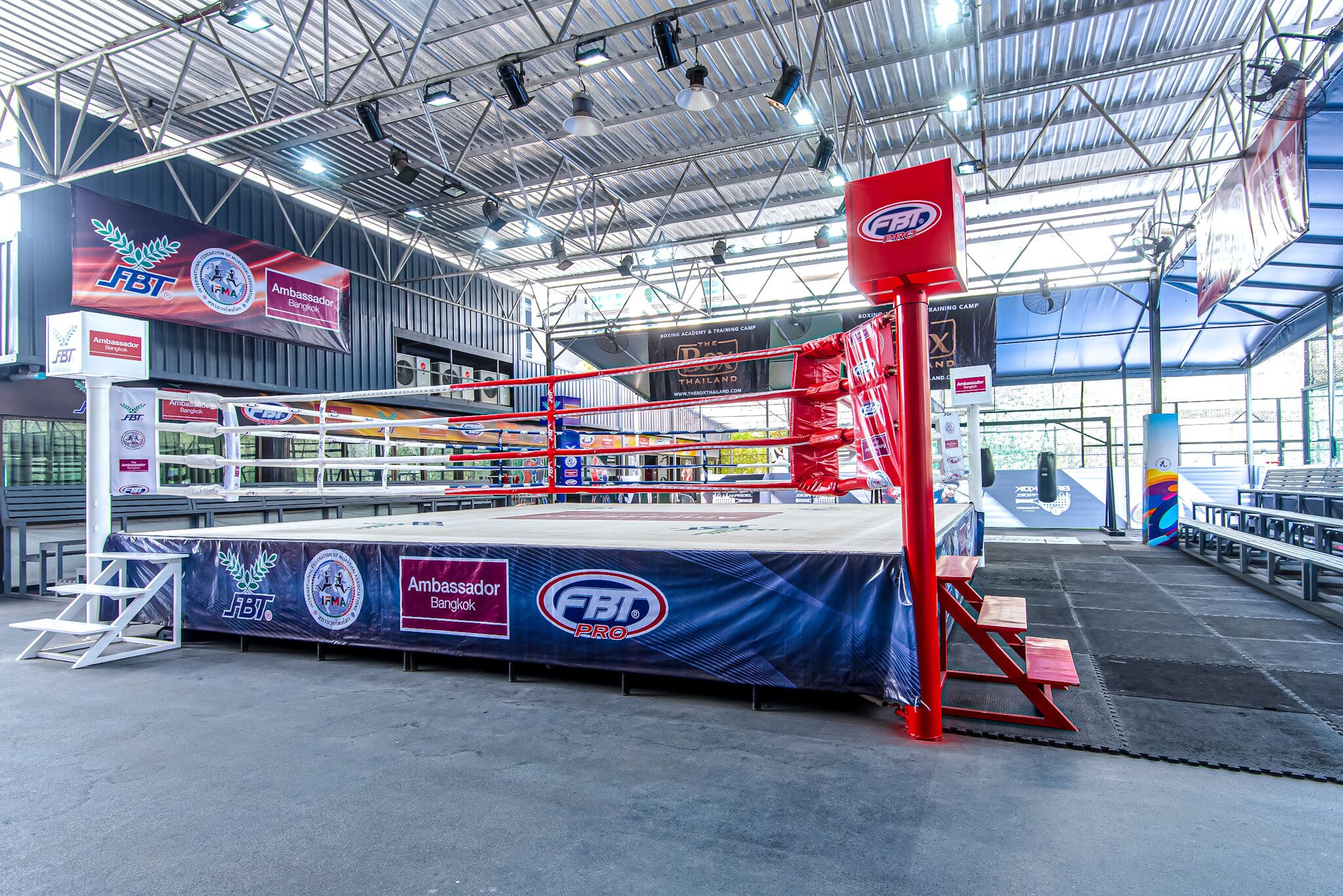 A boxing ring with a blue and red color scheme, surrounded by a metal fence, and various banners and logos are visible.