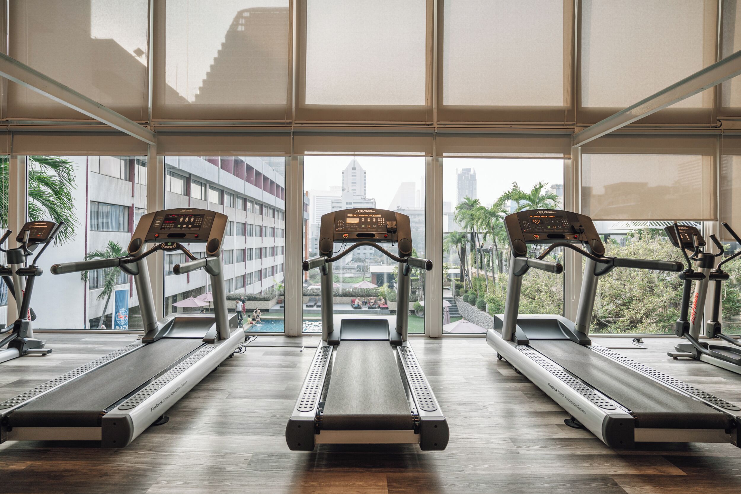 A gym with treadmills aligned in a row, large windows, and a view of buildings and palm trees outside.