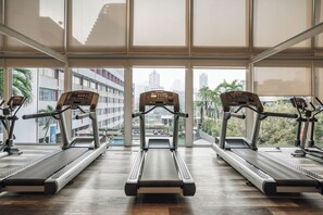 Un gimnasio con cintas de correr alineadas, amplias ventanas y vistas a edificios y palmeras.