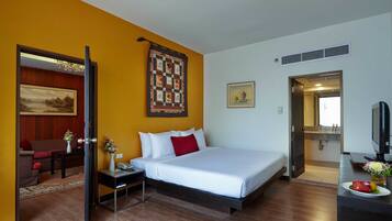 A hotel room with a bed, a desk, a chair, a small table with a vase, and a painting on the wall.