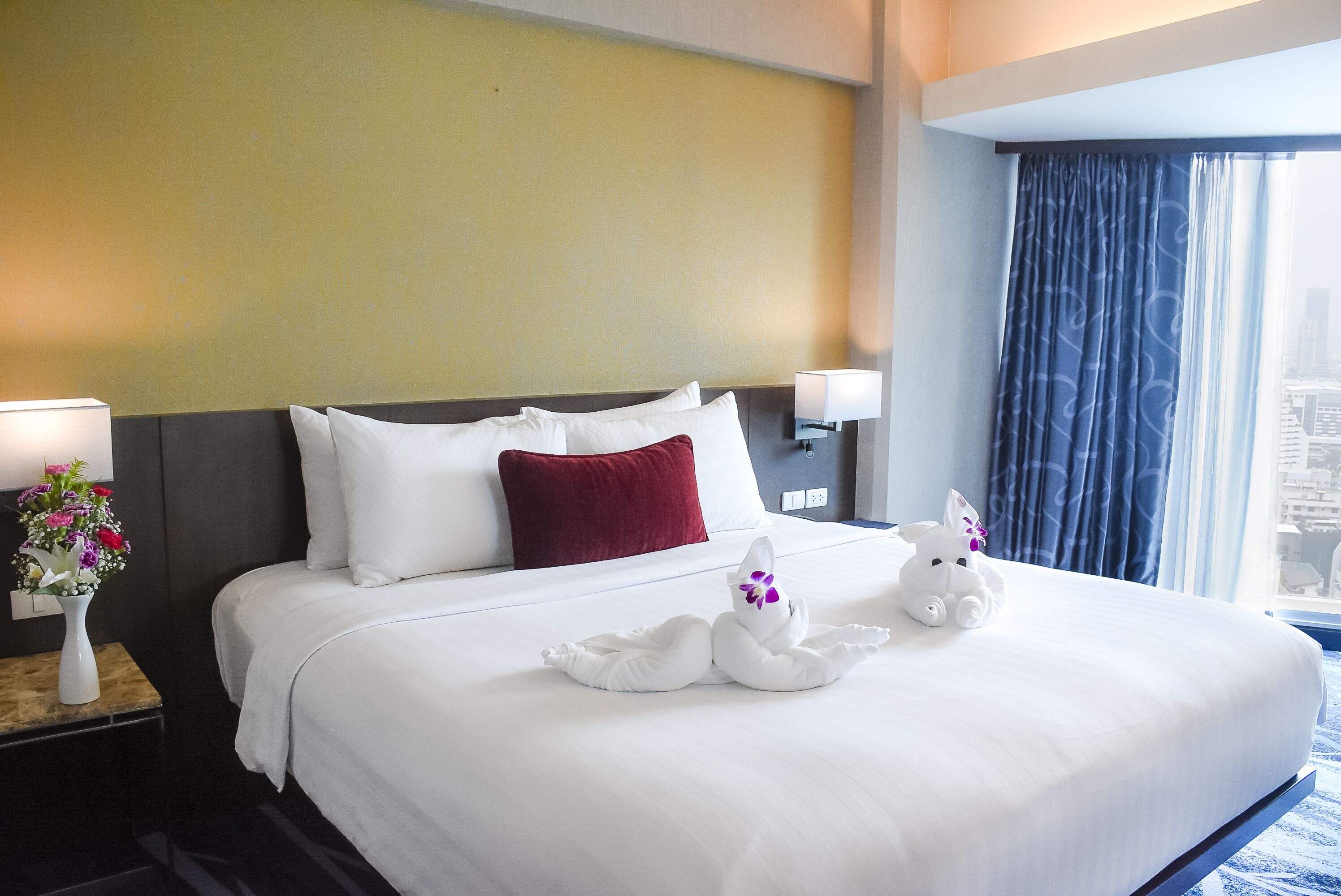Habitación de hotel con una cama grande, ropa blanca, una almohada roja, un florero con flores y una ventana con cortinas.
