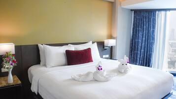 Habitación de hotel con una cama grande, ropa blanca, una almohada roja, un florero con flores y una ventana con cortinas.