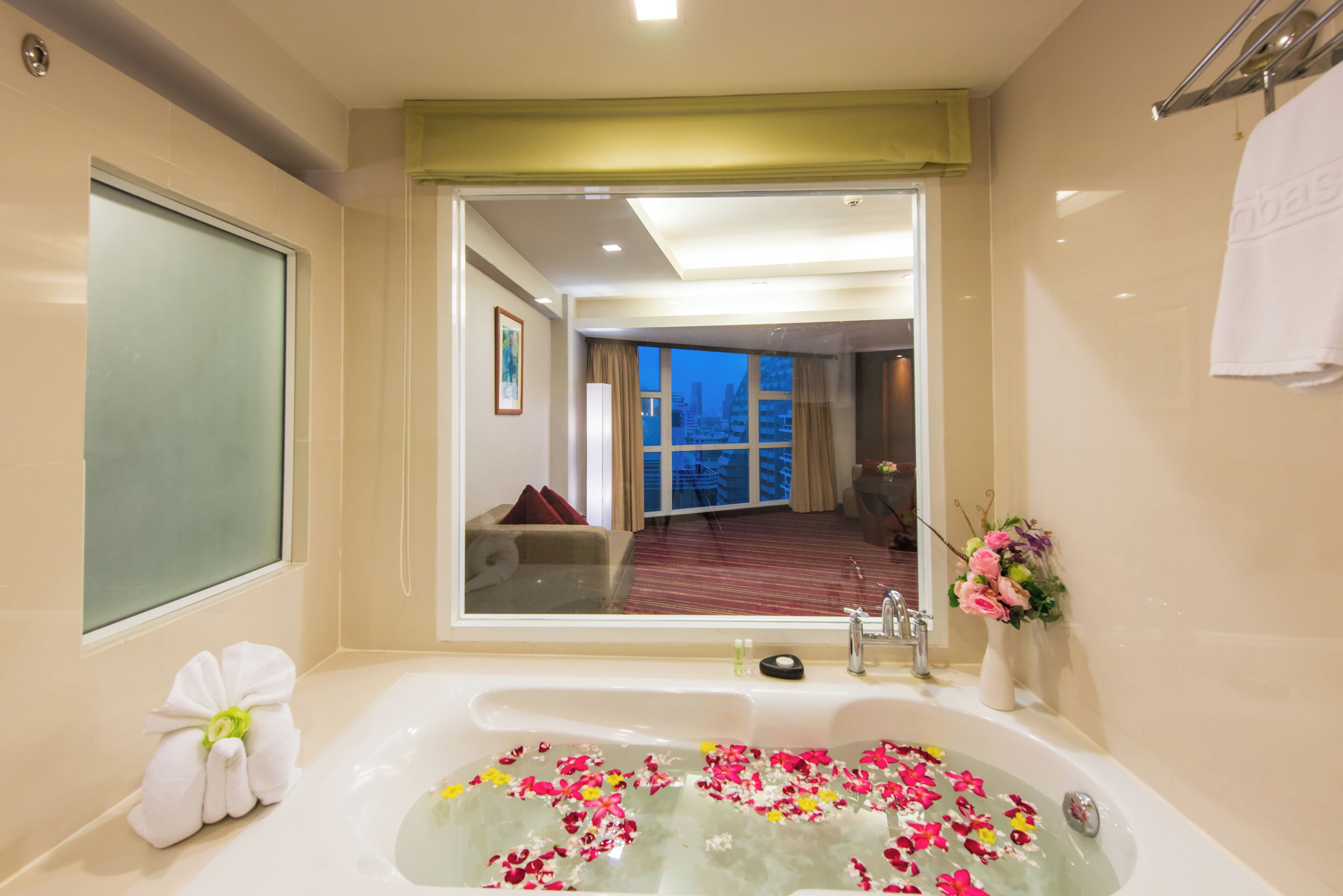A bathroom with a large bathtub filled with water and flower petals, a mirror, and a view of a living area through a glass door.