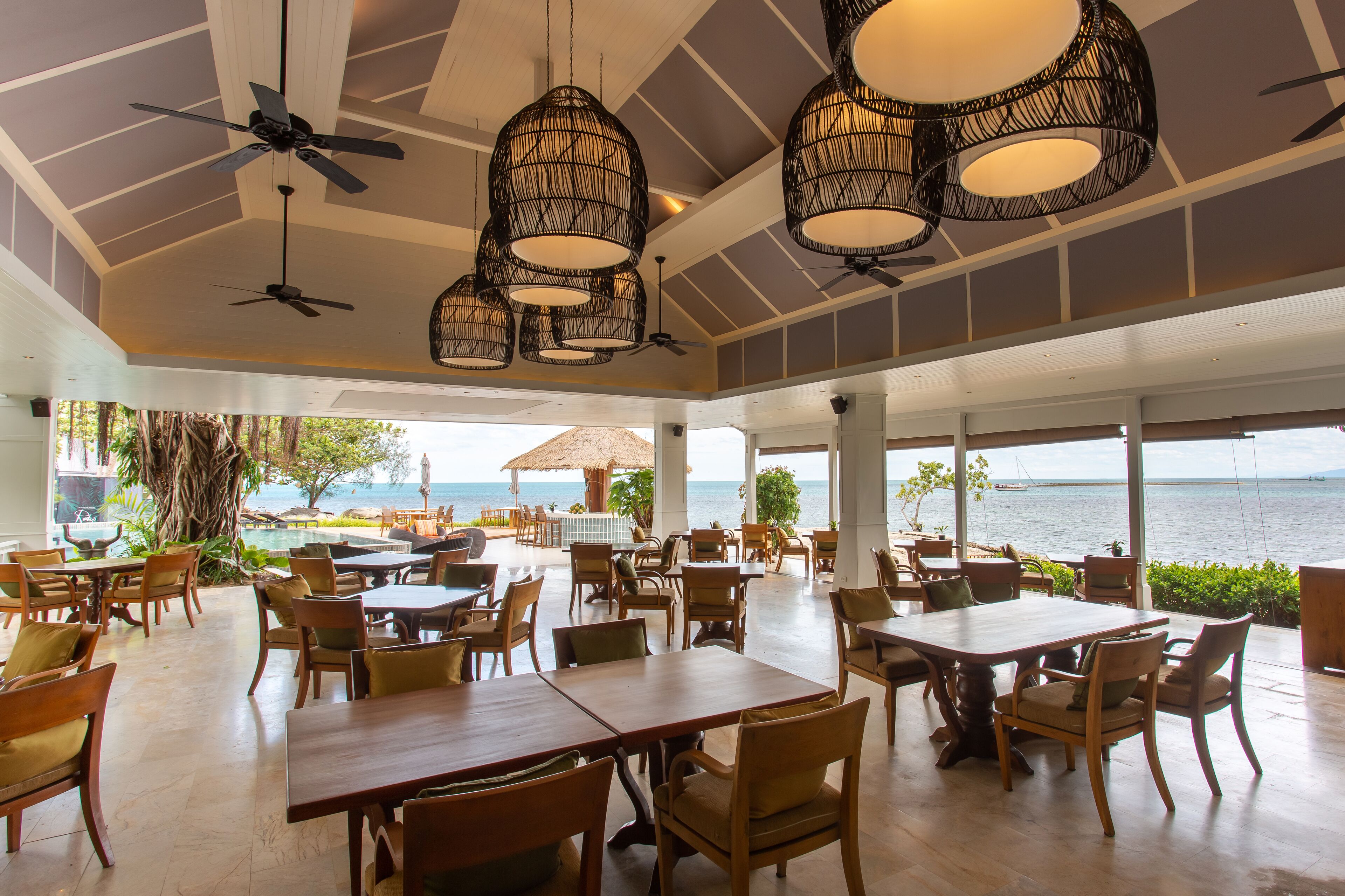 A spacious outdoor dining area with multiple tables and chairs, ceiling fans, and large windows offering a view of the sea.