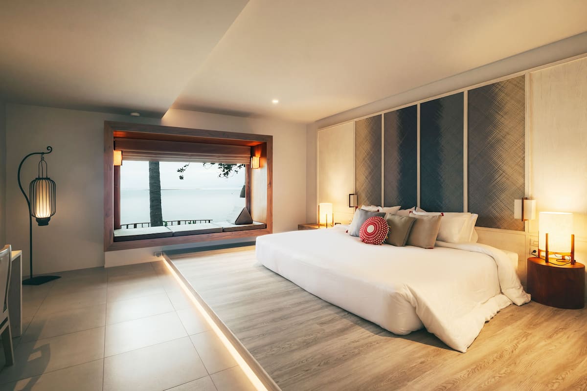 Une chambre moderne avec un grand lit, des lampes de chevet, une vue sur la mer et une fenêtre donnant sur des palmiers.
