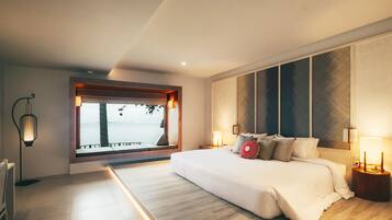 A modern bedroom with a large bed, bedside lamps, a view of the sea, and a window with a view of palm trees.