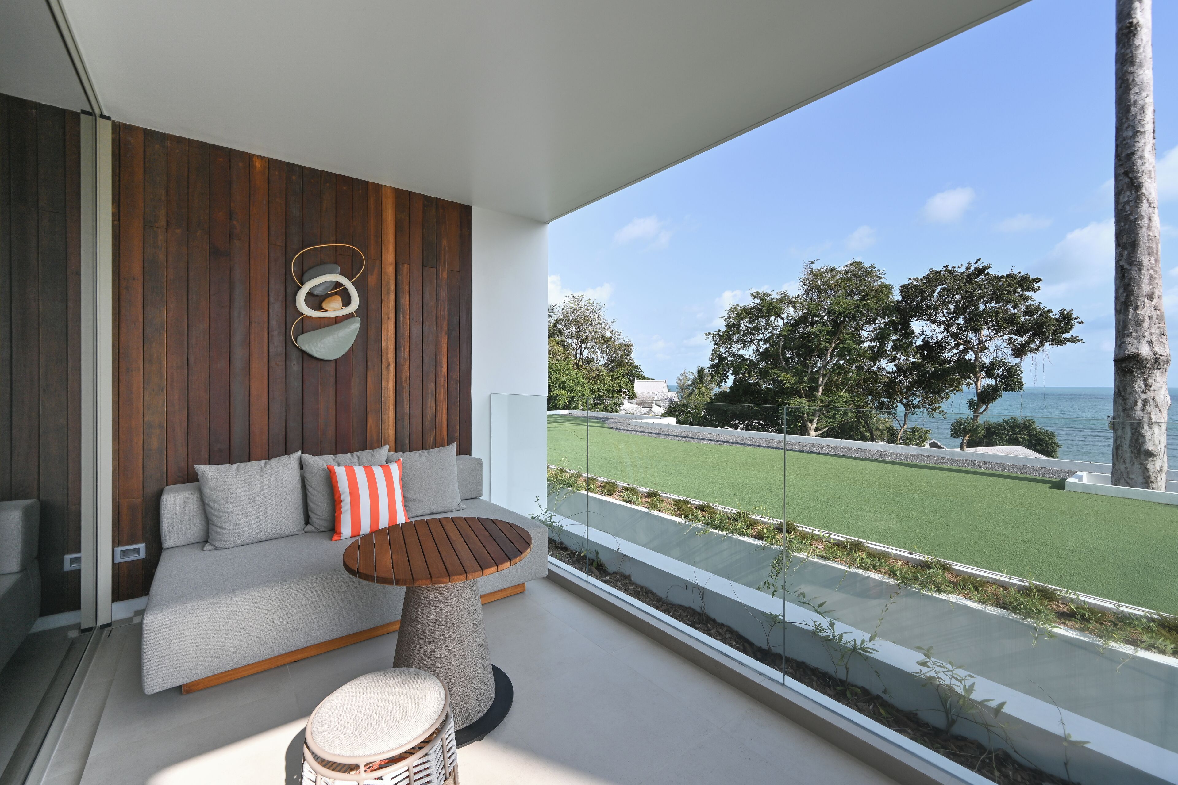 A modern balcony with a sofa, a round table, and a drum. It overlooks a green lawn and a body of water.