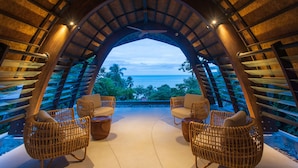 Un espacio de estar al aire libre con arco, sillas de mimbre y ventilador de techo, con vistas a un paisaje marino y vegetación exuberante.