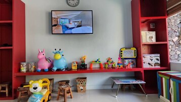 Un espace de jeux pour enfants avec des jouets, un téléviseur diffusant une image d’une pièce, et une bibliothèque contenant des livres et une horloge.