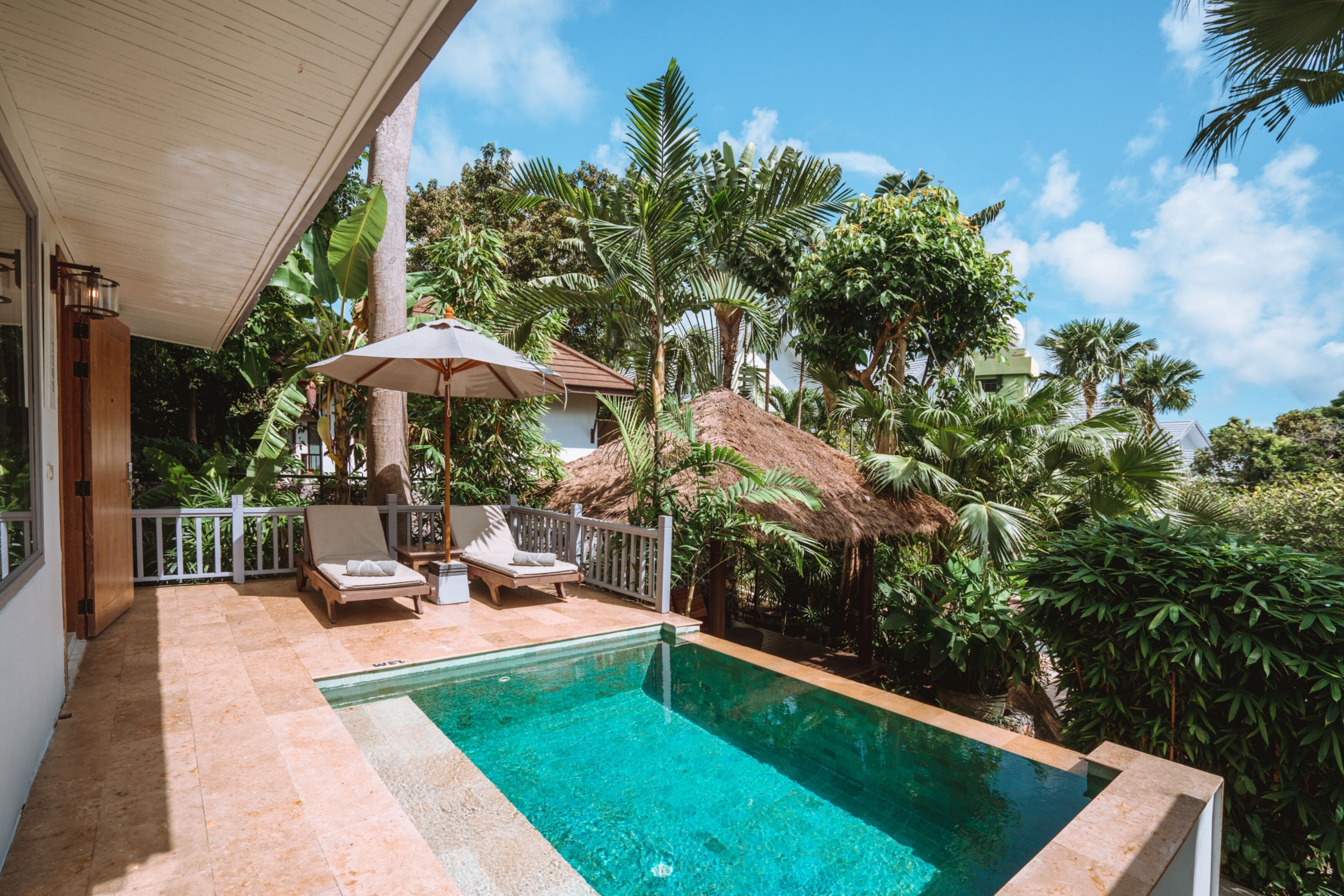 A swimming pool with lounge chairs and a thatched-roof structure surrounded by lush greenery.