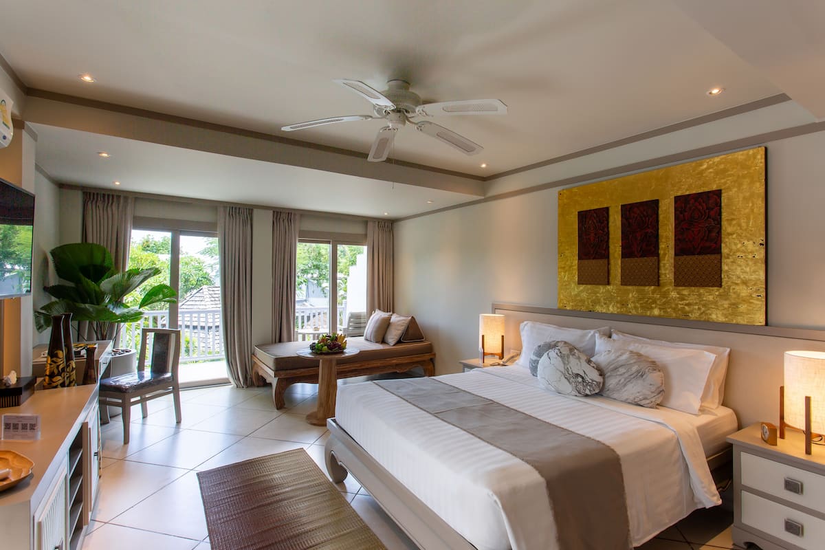 Une chambre spacieuse avec un grand lit, un ventilateur de plafond, un bureau avec une chaise et une fenêtre avec des rideaux.