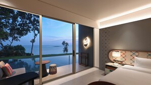 Habitación de hotel moderna con un gran ventanal corredero que da a un balcón con vistas al océano y palmeras. La habitación tiene una cama, una mesita de noche, una lámpara y una silla de mimbre.