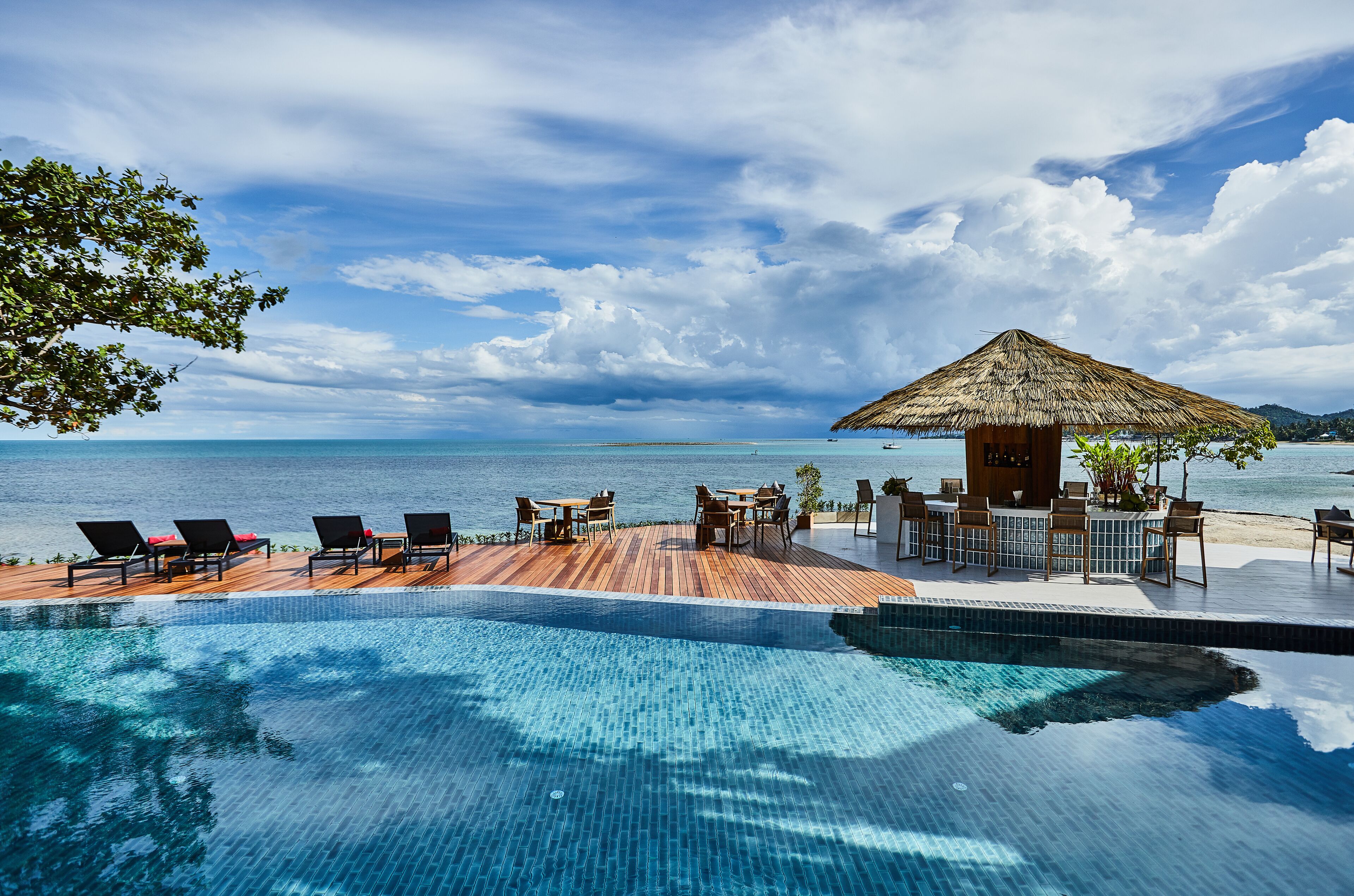 A poolside area with lounge chairs and tables, a thatched-roof structure, and a view of the sea.