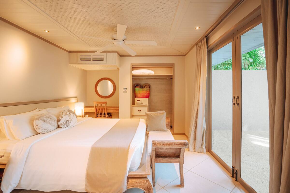 Une chambre d’hôtel avec un grand lit, un ventilateur de plafond, une petite table, une chaise, une armoire et une porte-fenêtre coulissante donnant sur l’extérieur.