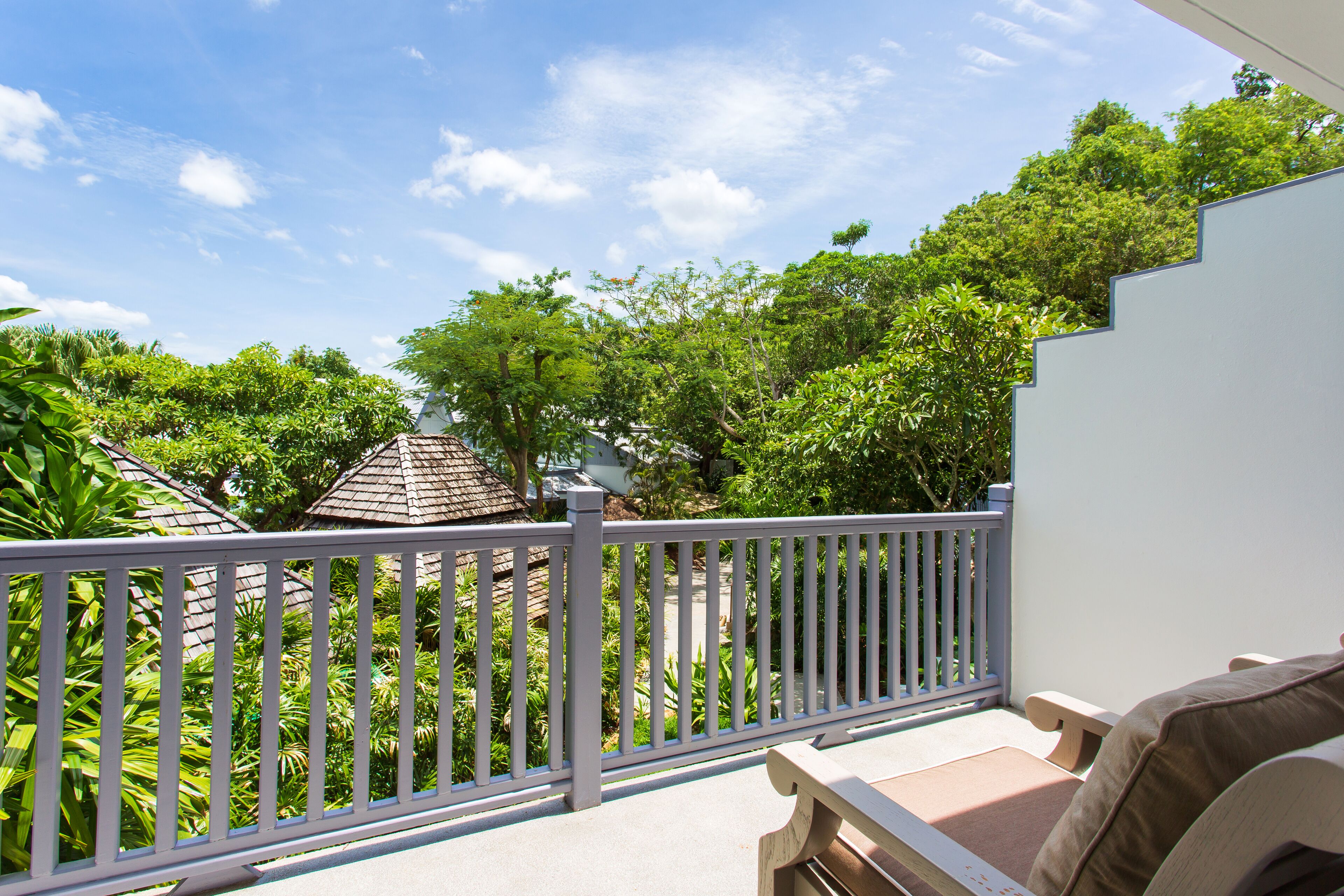 A balcony with a white railing, a bench, and a view of lush greenery and a building with a tiled roof.