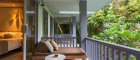 A balcony with a sofa and a table, a bed with pillows, and a view of lush greenery.