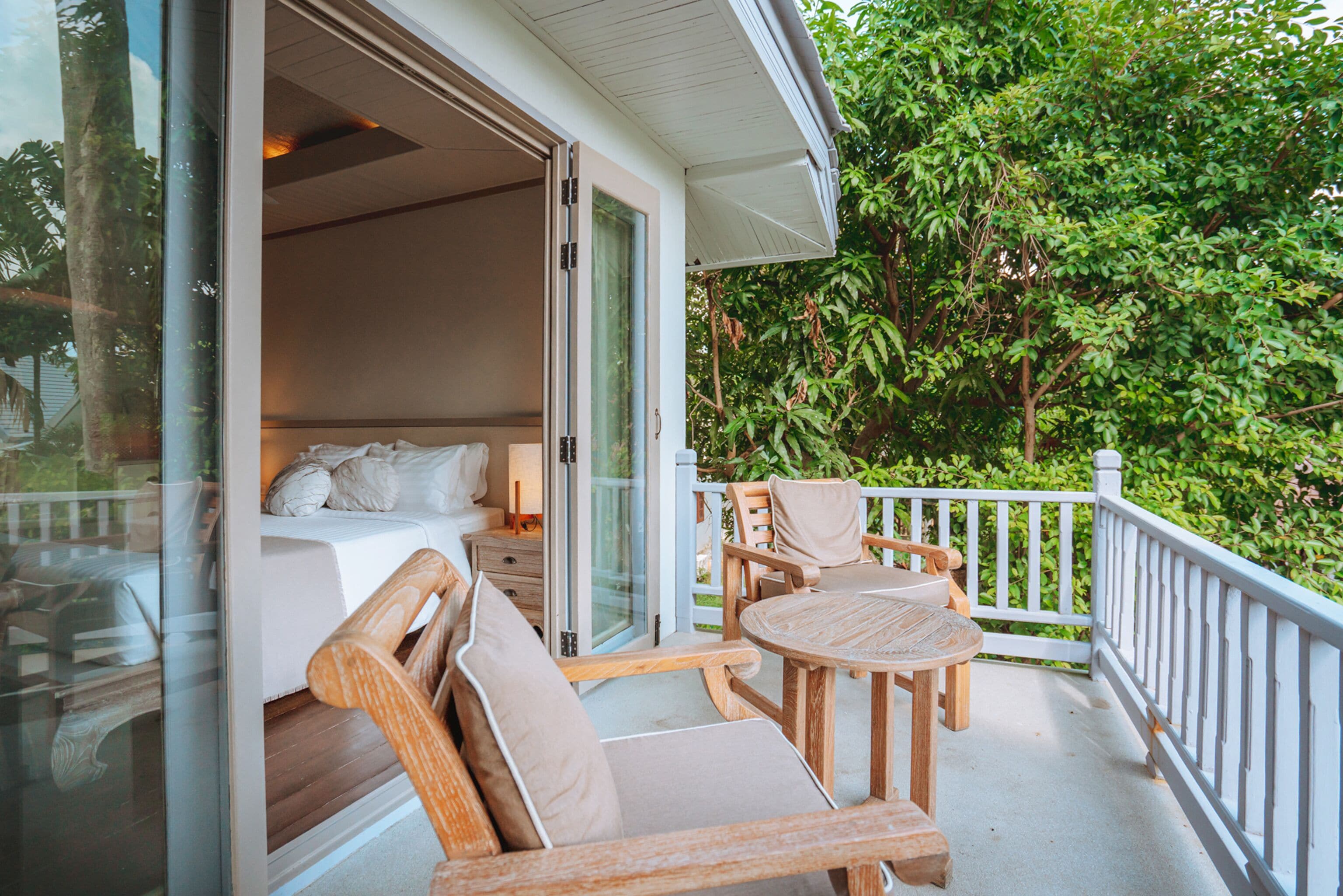 Een balkon met een houten stoel en tafel, een bed met witte beddengoed, en uitzicht op weelderige begroeiing.