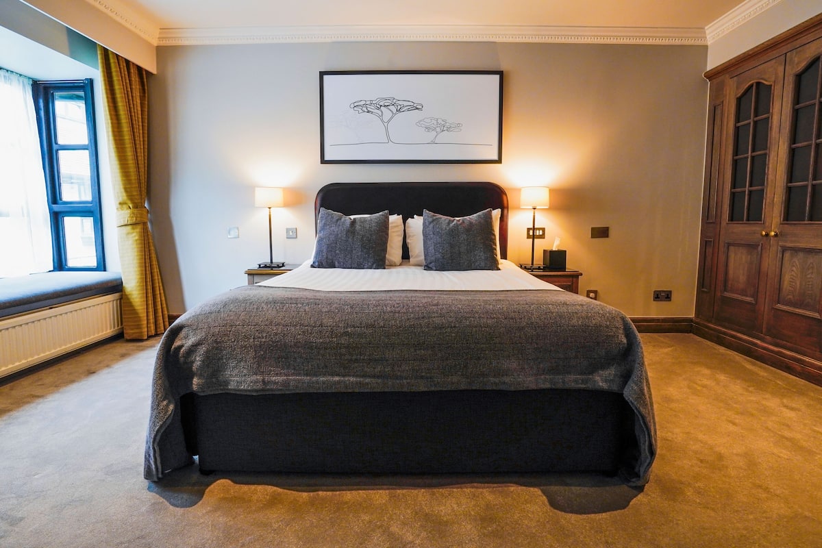 A neatly arranged bedroom with a large bed, two bedside lamps, a window with curtains, a radiator, and a wooden wardrobe.
