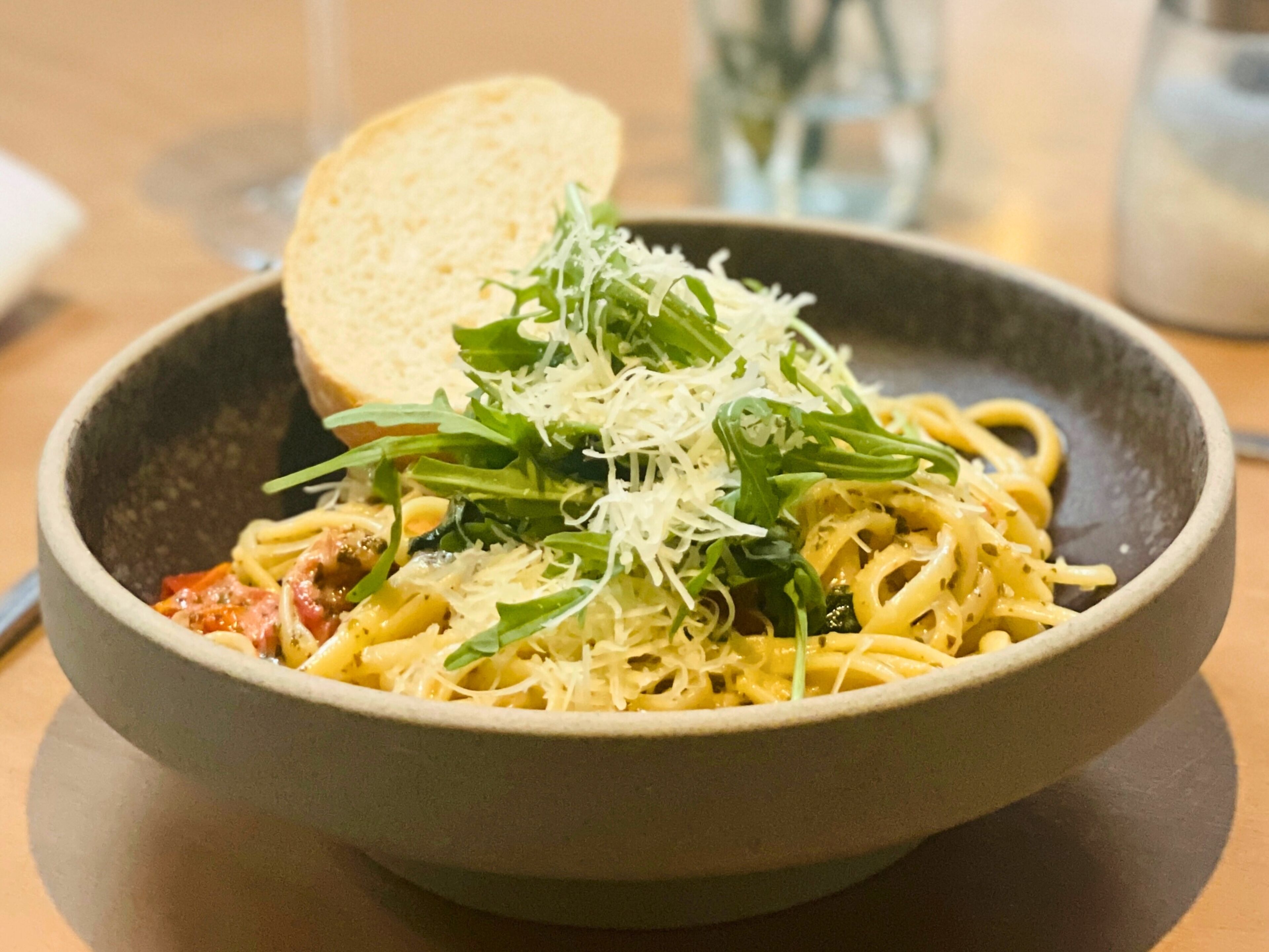 A bowl of pasta with greens and cheese, a slice of bread, and a glass of water on a table.