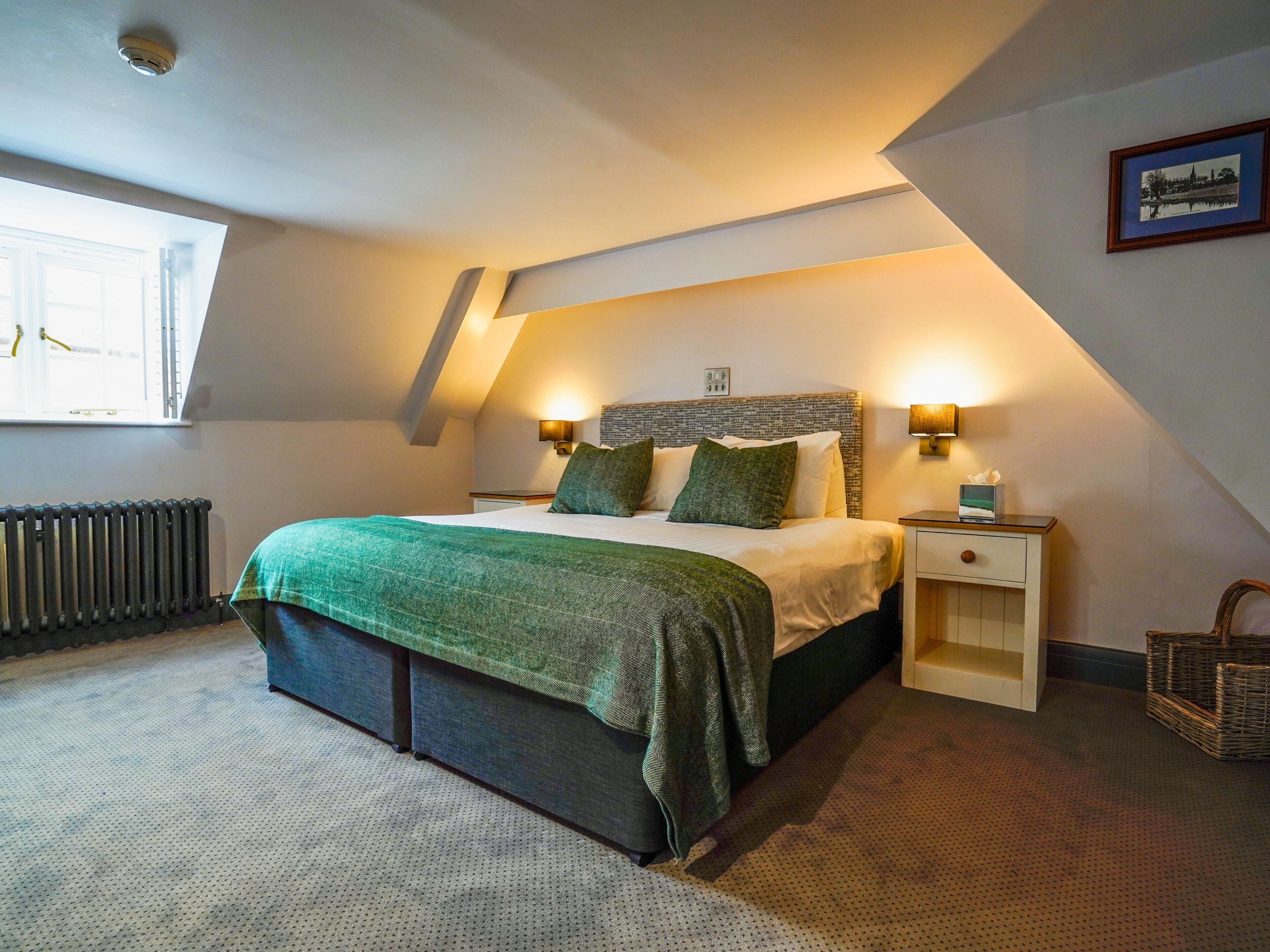 A bedroom with a bed, two bedside tables, a nightstand, a radiator, a window, and a framed picture on the wall.