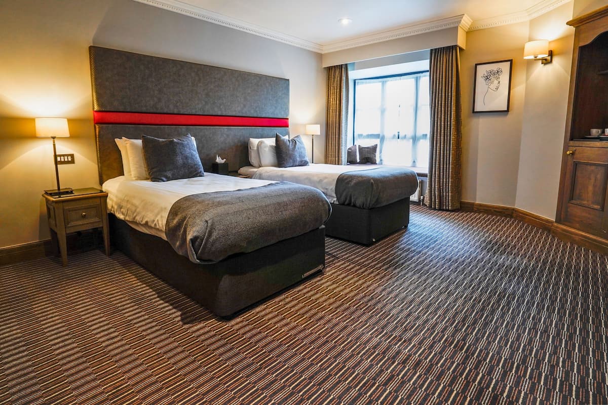A hotel room with a large bed, two bedside tables, a red accent on the headboard, a window with curtains, and a carpeted floor.