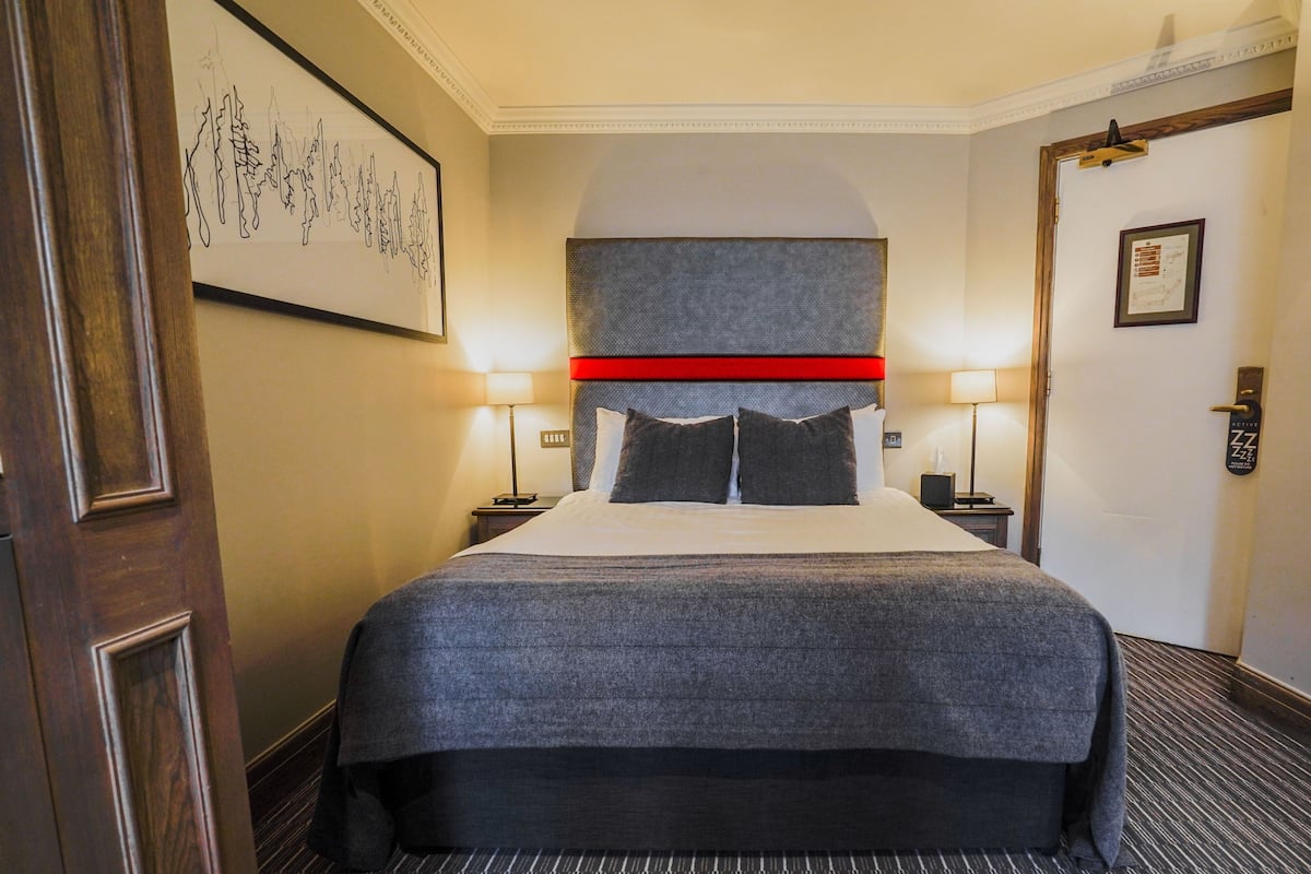 A hotel room with a large bed, two bedside tables, a headboard, a framed picture on the wall, and a door with a sign.