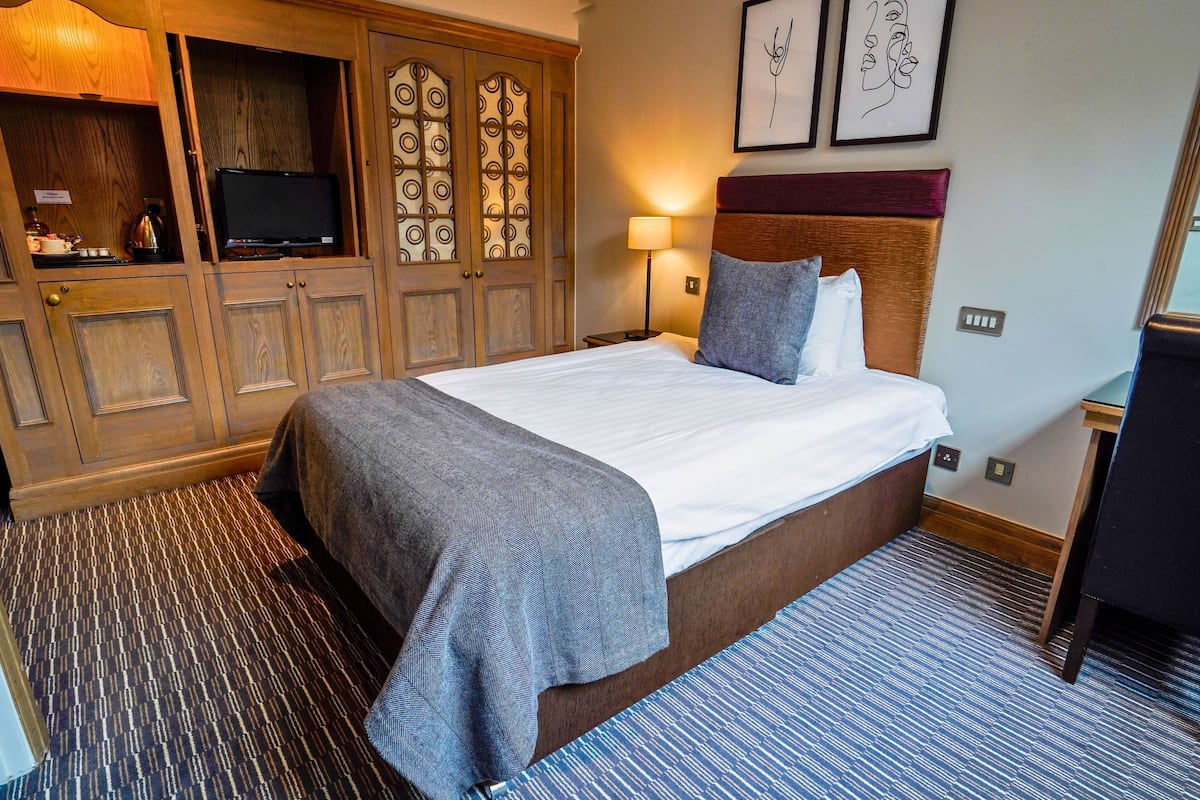 A hotel room with a large bed, wooden wardrobe, a television, and artwork on the wall.