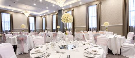 A banquet hall with round tables set for dining, chandeliers, and floral centerpieces.