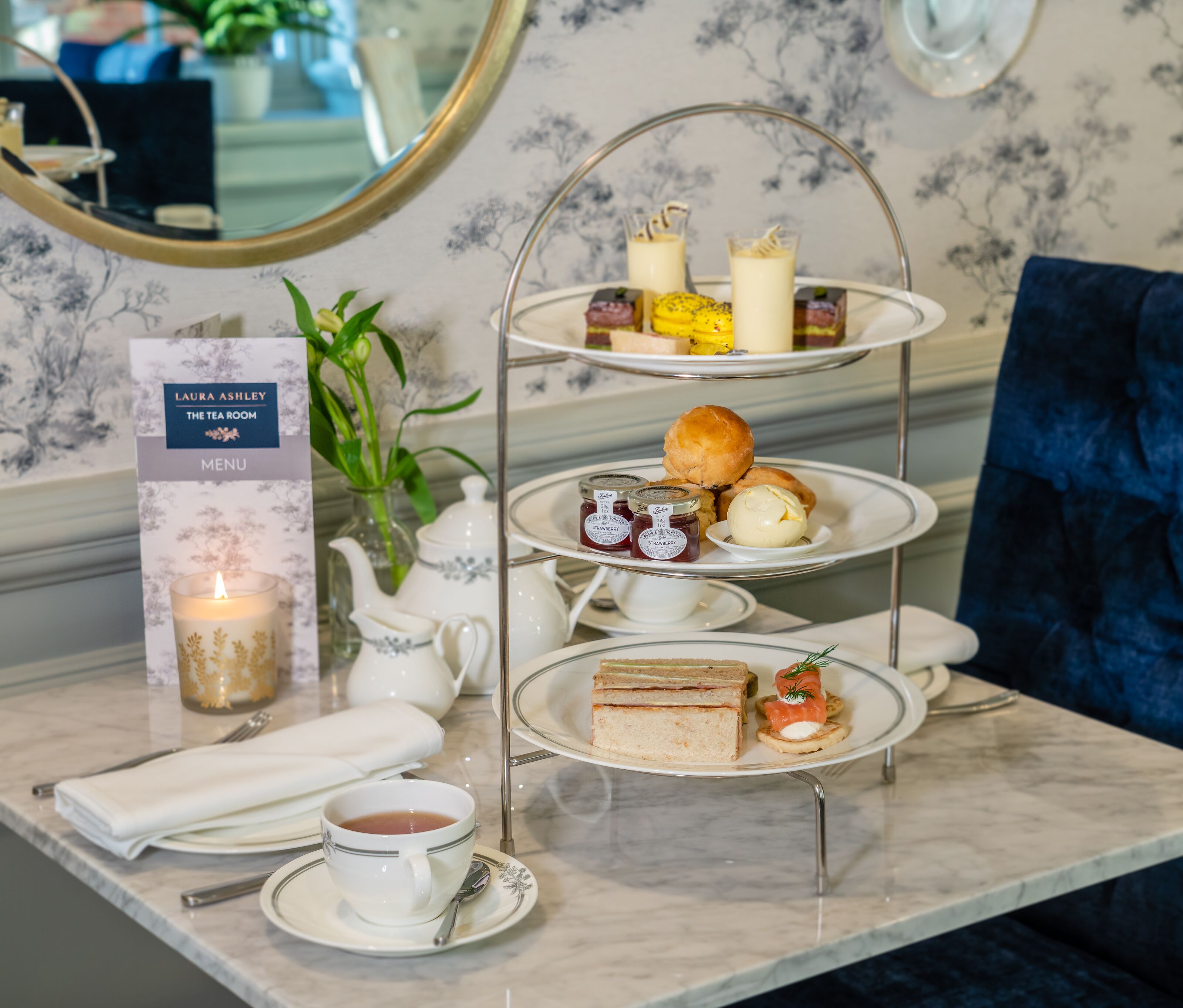 Un service à thé à trois niveaux, posé sur une table en marbre, accompagné d’un menu, d’une théière et d’une bougie.