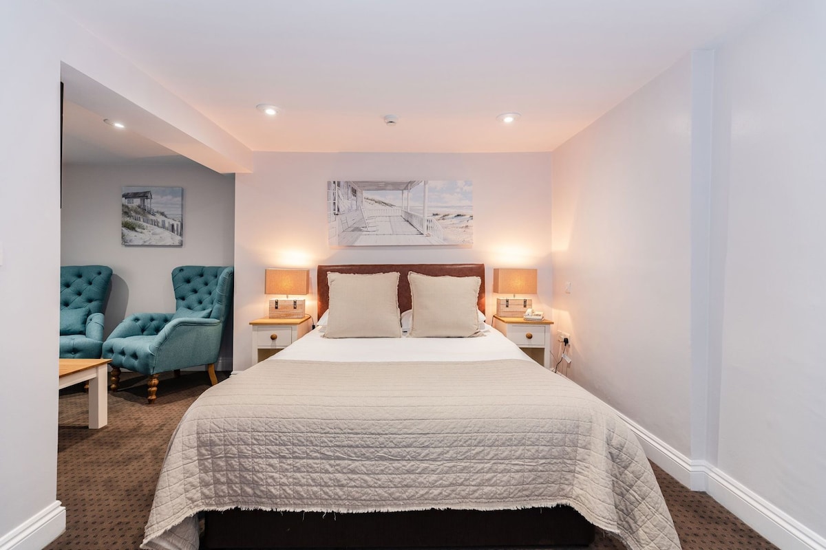 A bedroom with a bed, two nightstands, a headboard, a wall-mounted artwork, and two upholstered chairs.