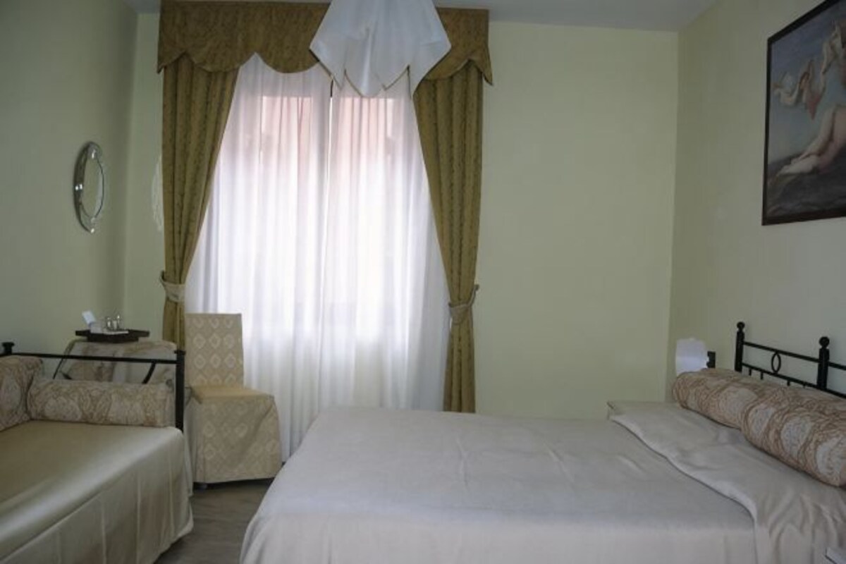 A hotel room with a bed, a chair, a bedside table, and a window with curtains.