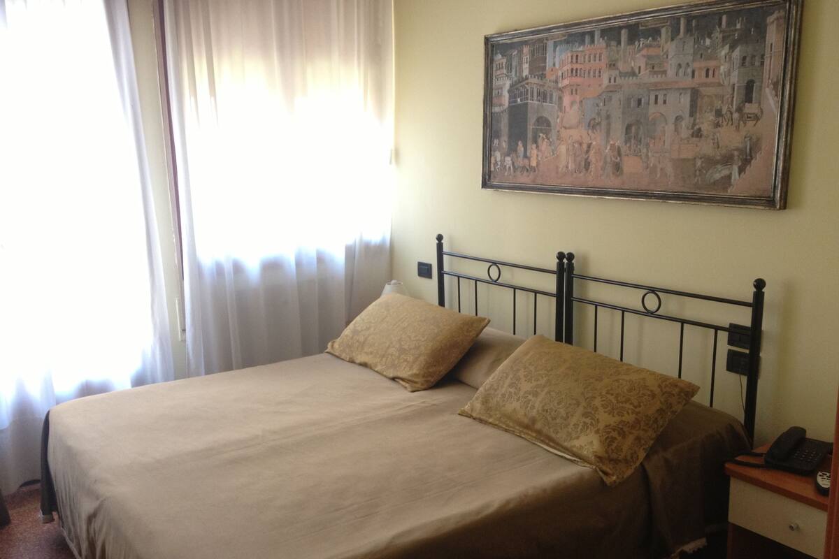 A hotel room with a bed, a nightstand, a painting on the wall, and a window with curtains.