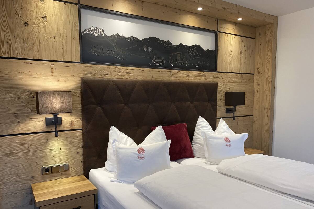 A hotel room with a wooden headboard, a mounted picture of a mountain landscape, a bedside table with a lamp, and two beds with white bedding and decorative pillows.