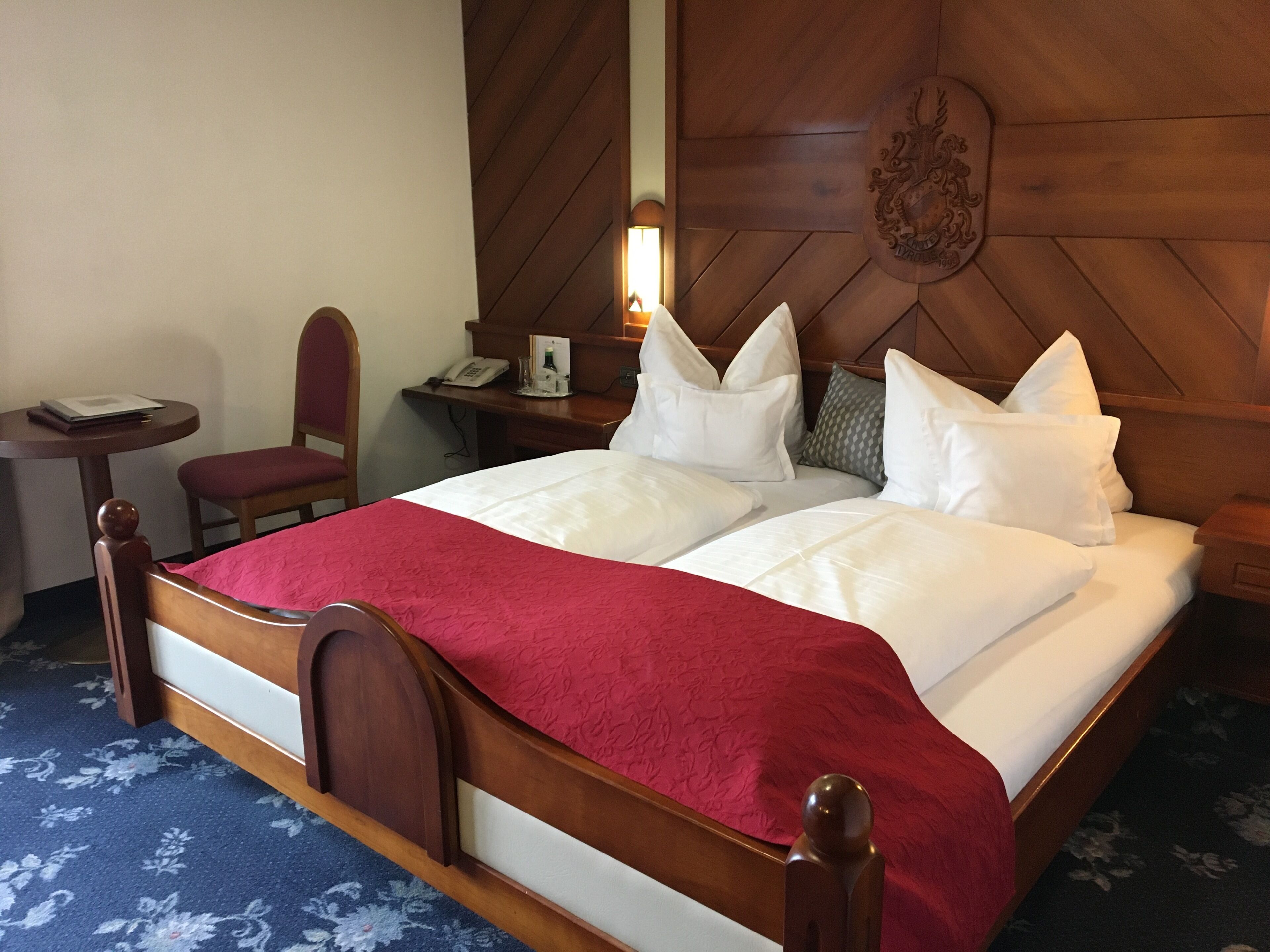 A hotel room with a wooden headboard, a bed with white linens and a red throw, a desk with a telephone, and a chair.