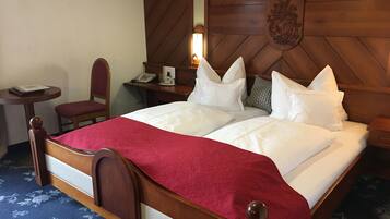 A hotel room with a wooden headboard, a bed with white linens and a red throw, a desk with a telephone, and a chair.