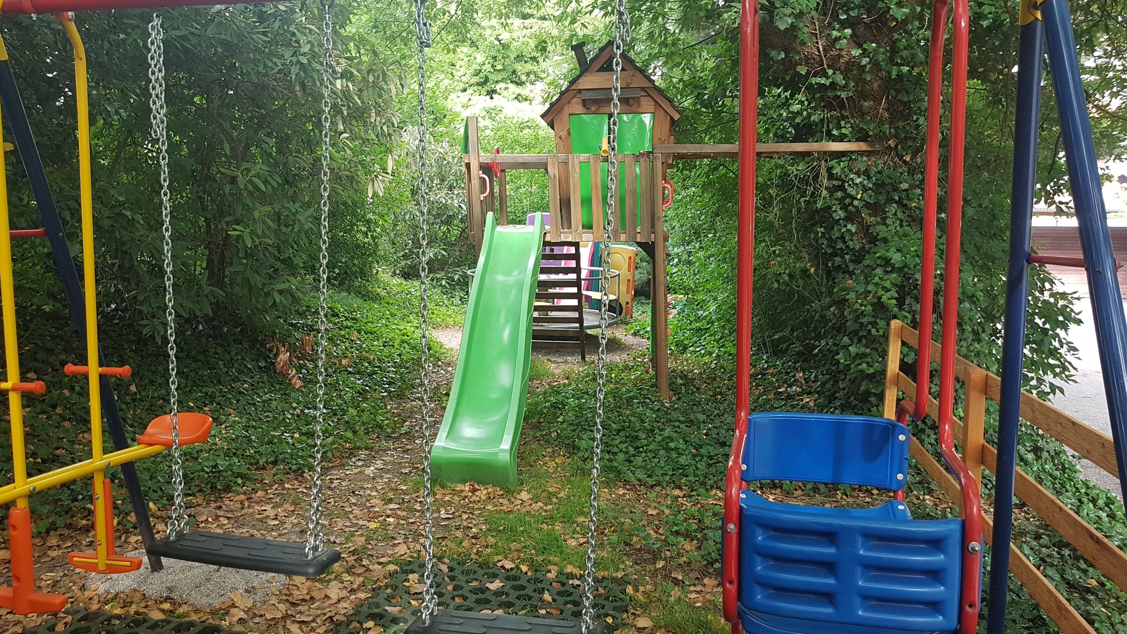 Ein Spielplatz mit Schaukeln, einer Rutsche und einer Kletterkonstruktion, umgeben von viel Grün.