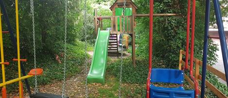 Un parque infantil con columpios, un tobogán y una estructura para escalar, rodeado de follaje.