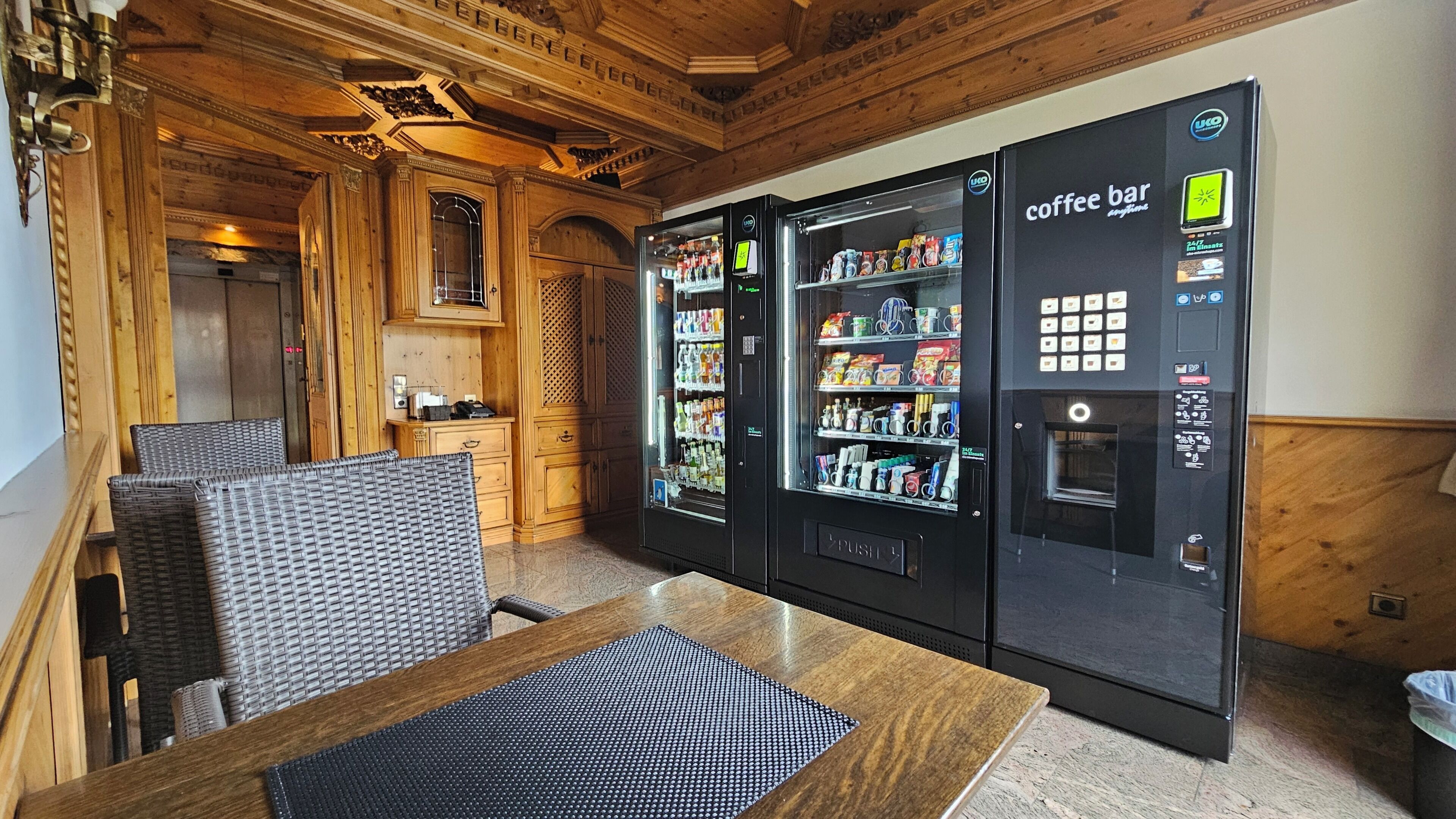 A coffee bar area with two vending machines, wicker chairs, a wooden table, and a wooden ceiling.