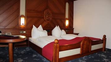 Habitación de hotel con cabecera de madera, mesita de noche redonda, cama con ropa blanca y dos lámparas fijadas a la pared.