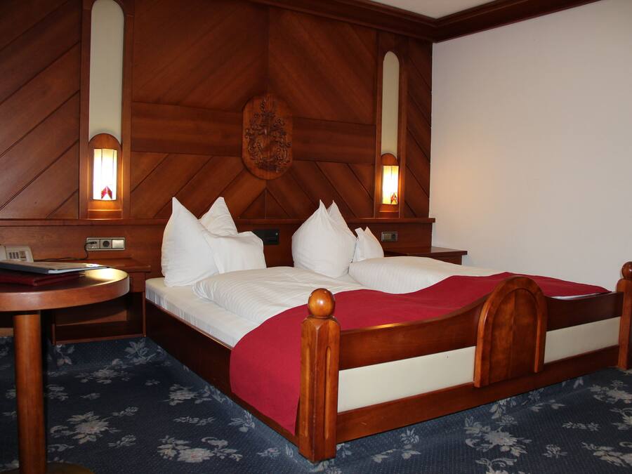 Habitación de hotel con cabecera de madera, mesita de noche redonda, cama con ropa blanca y dos lámparas fijadas a la pared.