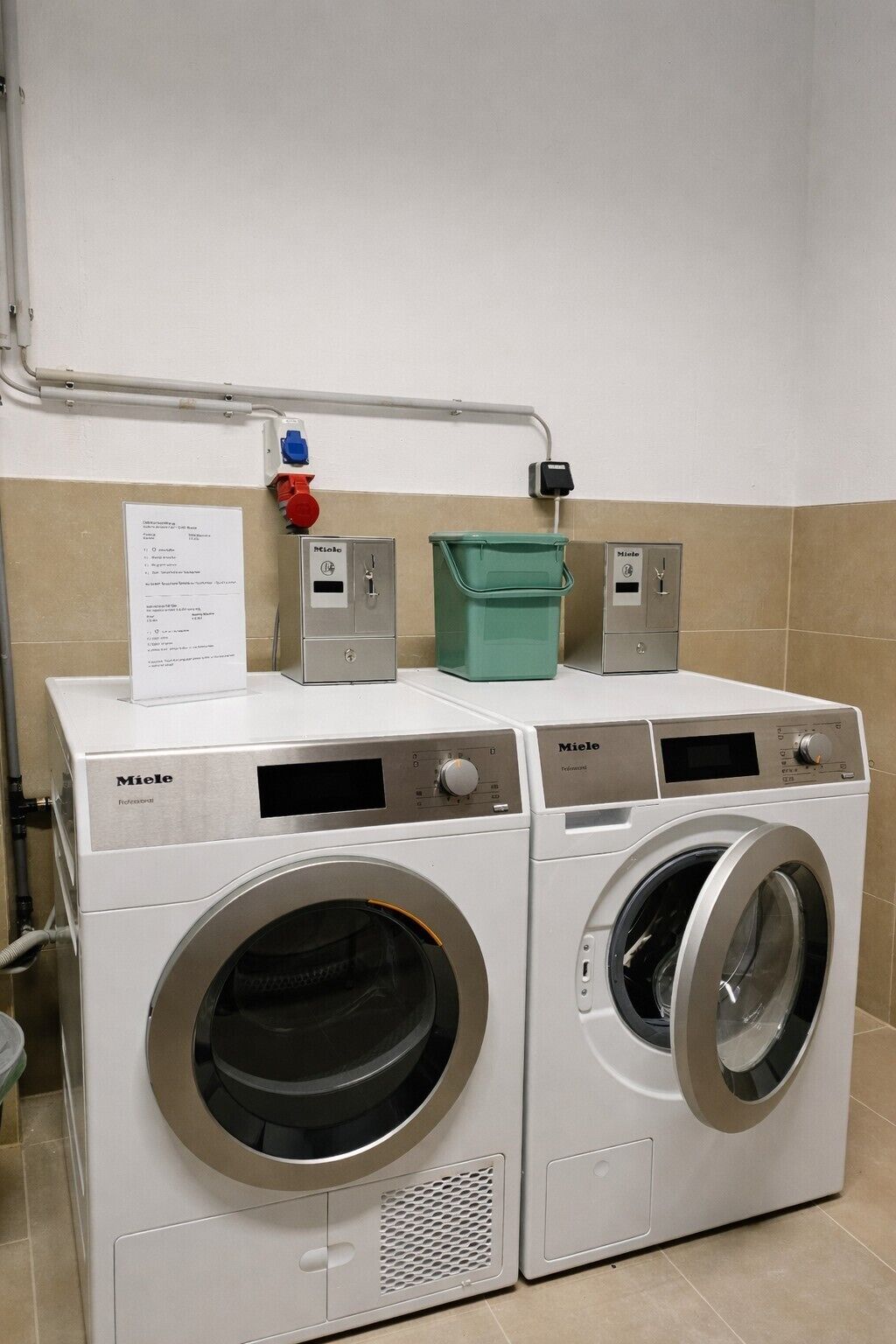 Two white Miele Professional front-loading washing machines in a laundry room with a beige and white tiled wall, a control panel, and a green laundry basket.