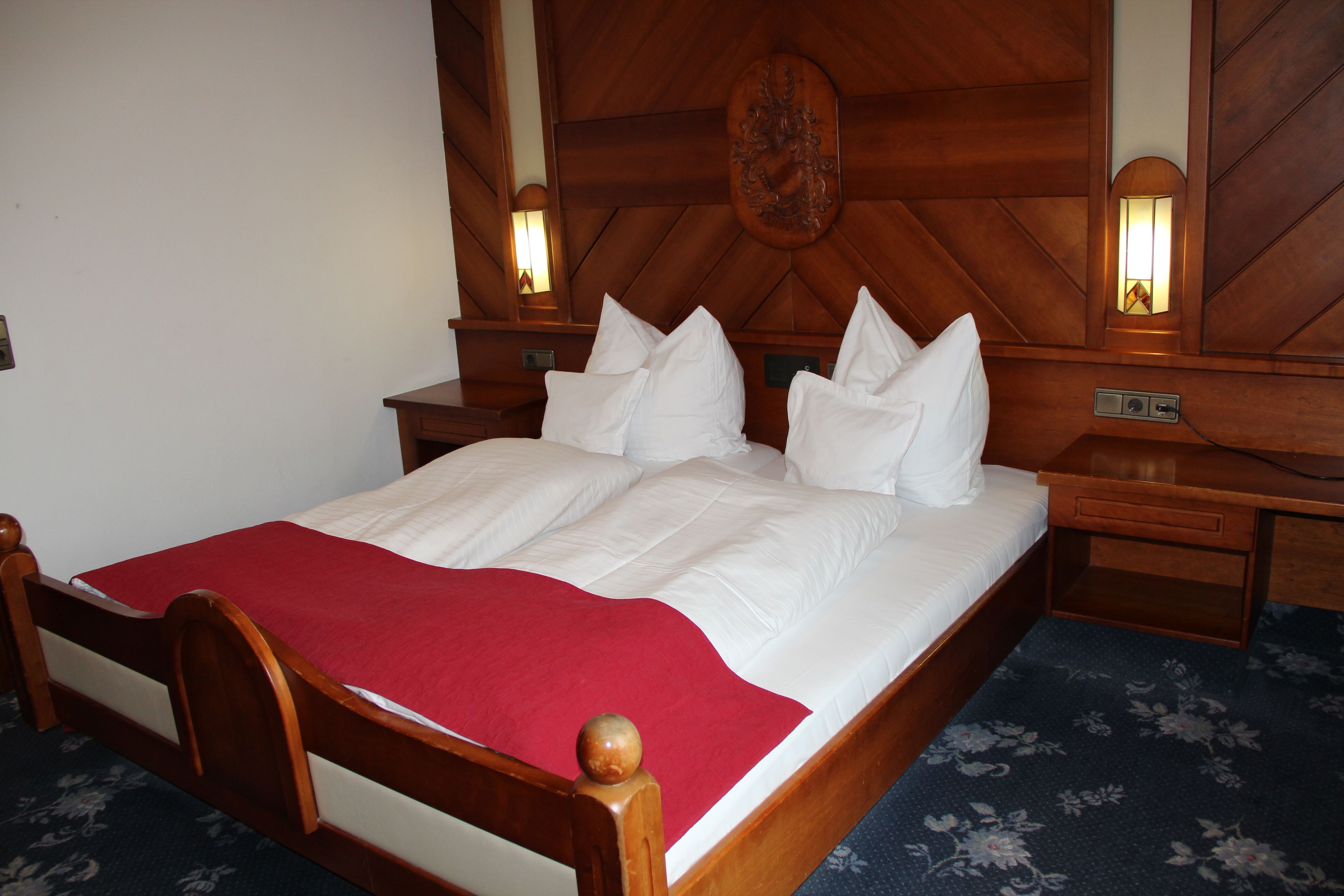 Una cama bien tendida con sábanas blancas y una colcha roja, cabecera de madera, dos mesitas de noche con lámparas y una alfombra estampada con motivos florales.