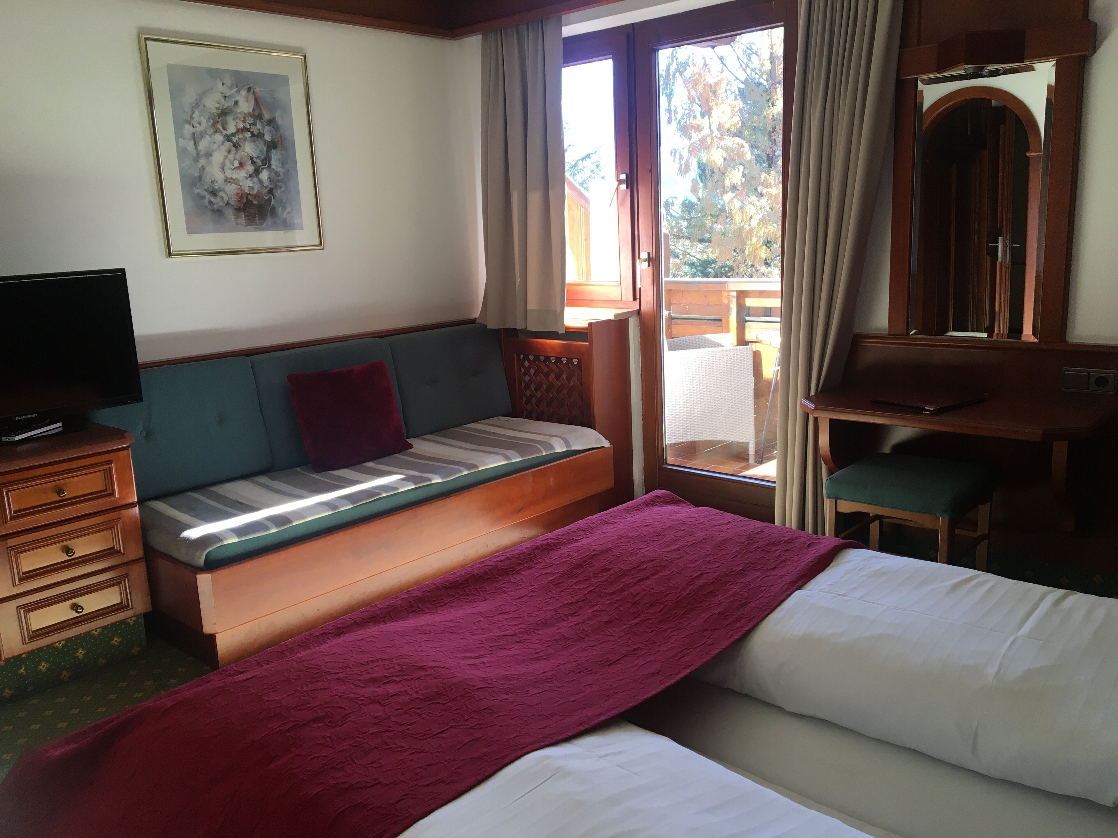Quarto de hotel com cama, sofá, escrivaninha, televisão e janela com cortinas.