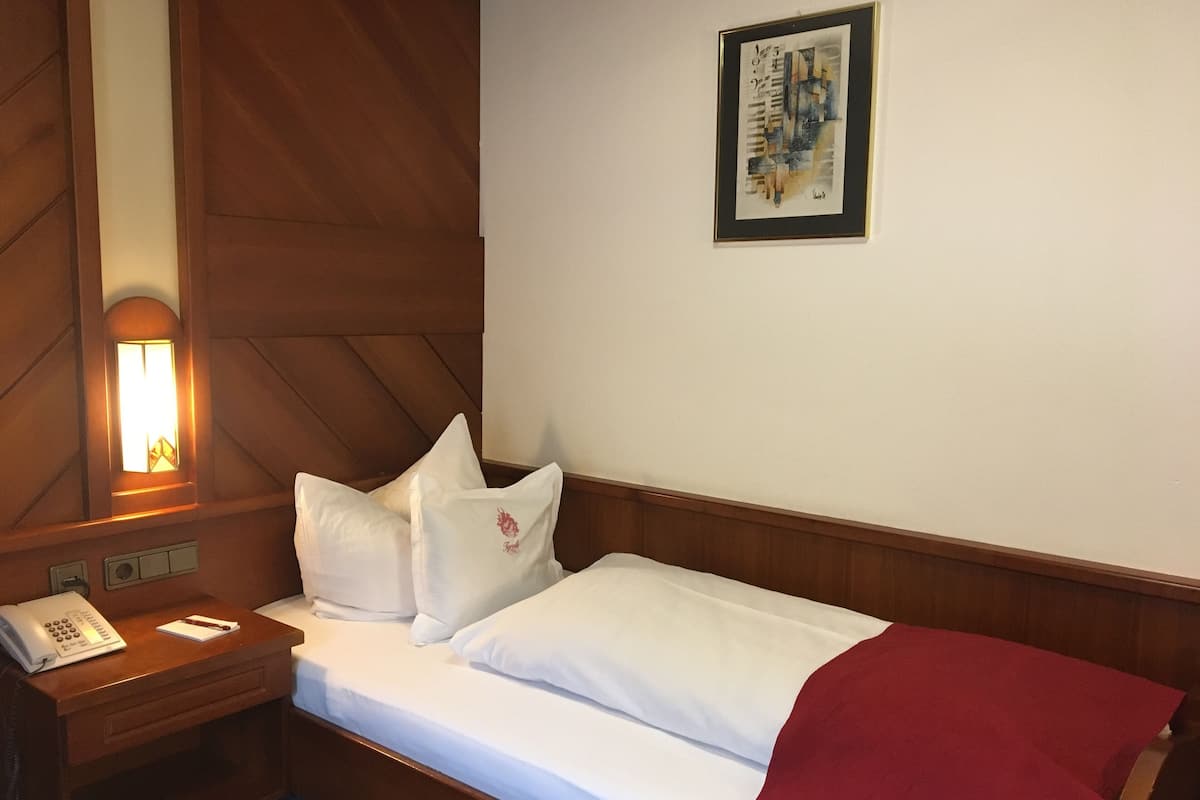 A hotel room with a wooden headboard, a bedside table with a phone and notepad, a red throw blanket, and a framed artwork on the wall.