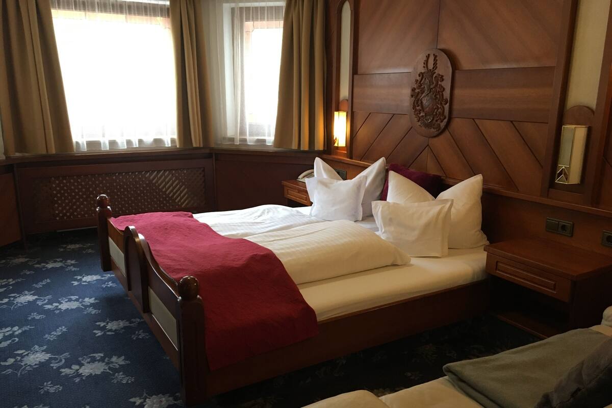 A hotel room with a wooden bed, a red throw blanket, white pillows, a nightstand, a mirror, and a decorative wall piece.