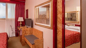 Quarto de hotel com cama, escrivaninha, cadeira, televisão e janela com cortinas.