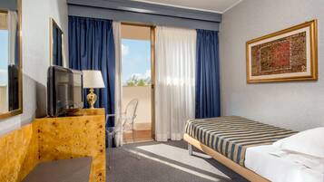 Quarto de hotel com uma cama, uma escrivaninha com computador, uma cadeira, uma televisão e uma varanda com cortinas.