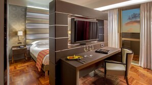 Una habitación de hotel moderna con cama, escritorio con televisor de pantalla plana, silla y ventana grande con cortinas.